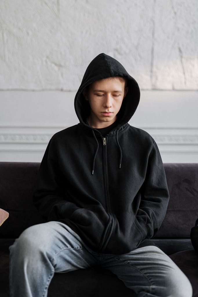 Teenager in a black hoodie sitting on a couch, reflecting on thoughts indoors.